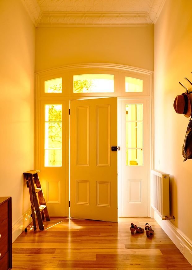 A warmly lit residential entryway featuring a classic paneled front door standing open, flanked by glazed sidelights and topped with fanlight-style transom windows. The space has polished timber flooring, ornate pressed-pattern ceiling panels, decorative cornices and wall mouldings, white trim, a wall-mounted radiator, a small timber ladder, and coats and shoes near the entrance. Strong golden sunlight creates a welcoming, historic character.