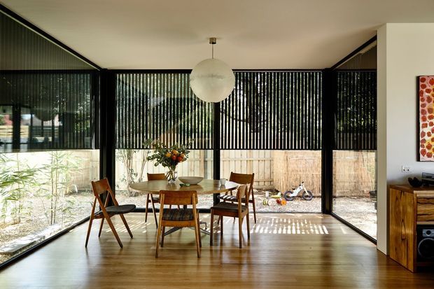 A bright, mid-century-inspired dining area enclosed by extensive black-framed glazing with vertical blinds. A timber dining table and assorted wooden chairs sit beneath a spherical pendant light, with a timber floor, built-in timber storage and a garden/paved outdoor area visible beyond the windows.