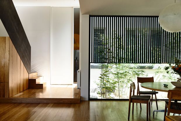 Warm contemporary open-plan interior featuring a timber-lined stair and raised landing, white built-in cabinetry, polished timber flooring, and a dining area beside full-height glazing with vertical black screening. Natural greenery is visible outside, and a large spherical pendant hangs over the dining space.