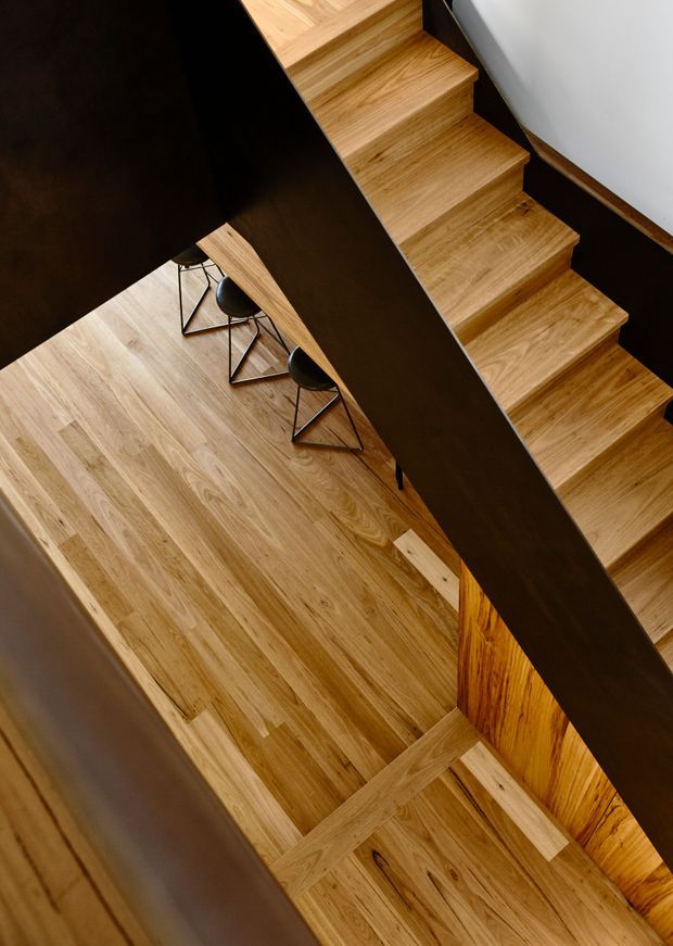 A high-angle interior view of a modern timber staircase crossing above a warm oak-finished floor. The stair treads and surrounding flooring are light to mid-toned timber, with strong dark structural balustrade or side panels creating bold diagonal lines. A small group of black stools is visible beneath the stair, and a timber-clad vertical feature adds warmth to the stairwell.