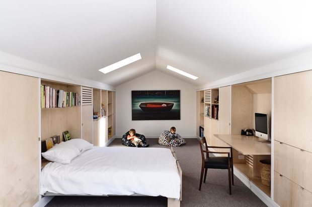A compact multifunctional room with a built-in wall bed in the foreground, extensive pale timber cabinetry and open shelving along both side walls, and a built-in desk with computer at right. Two people sit on the carpeted floor beneath a framed artwork. The room has a simple white pitched ceiling with slim linear skylights or ceiling lights, creating a bright, highly integrated and space-efficient interior.
