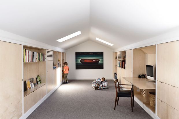 Bright, symmetrical multipurpose room with a pitched ceiling and extensive built-in pale timber cabinetry lining both side walls. The joinery incorporates open bookshelves, display niches, storage, and a built-in desk with computer. A carpeted central floor provides a flexible workspace or study area, with a framed artwork on the rear wall, a child, and a patterned floor cushion. The room has white walls and ceiling, integrated linear ceiling lights, and a clean contemporary built-in design.