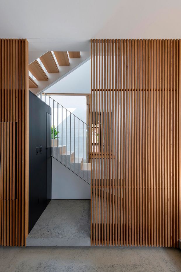 Interior view of a contemporary stairway and hallway framed by floor-to-ceiling vertical timber slat screens. A raw concrete floor and stair, dark painted wall, slim metal balustrade, and timber-clad ceiling beams create a warm, minimal architectural composition with filtered light and glimpses of greenery beyond.