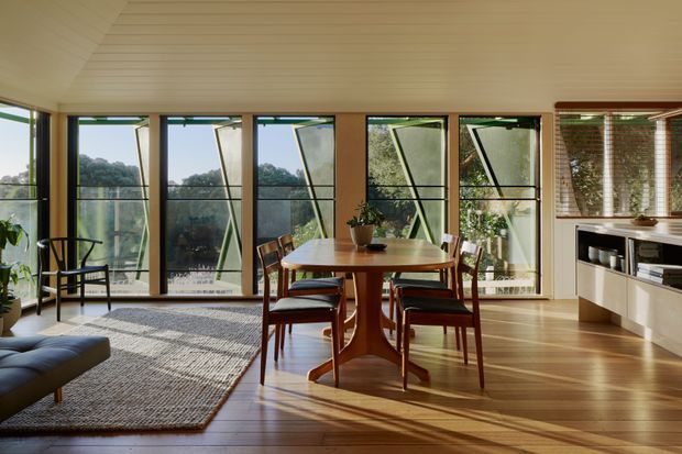 A warm, light-filled open-plan dining and living area with a mid-century-inspired timber dining table and upholstered chairs, a low sofa at left, and extensive floor-to-ceiling glazing overlooking greenery. The room features timber flooring, a white-lined sloped ceiling, slim dark window frames, and built-in shelving beneath timber venetian blinds.