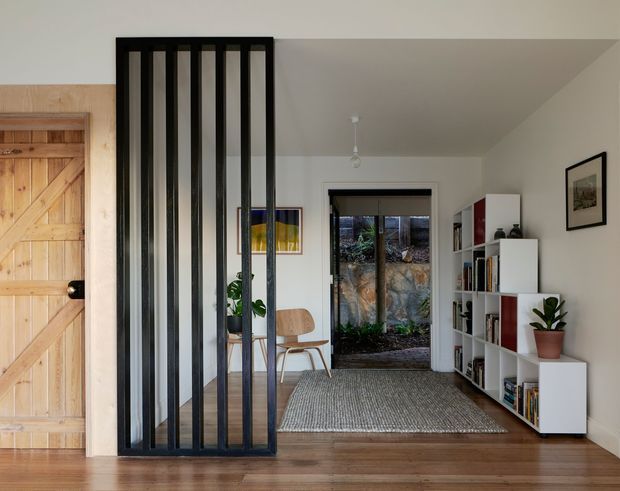 A bright, refined residential interior viewed from a timber-floored entry or hallway. A black vertical slatted room divider separates the entry from a small sitting area, with a natural timber barn-style door on the left, white walls, a low-pitched ceiling, built-in white shelving filled with books and objects, a woven grey rug, timber chair, indoor plant, and glazing opening toward a planted outdoor area.