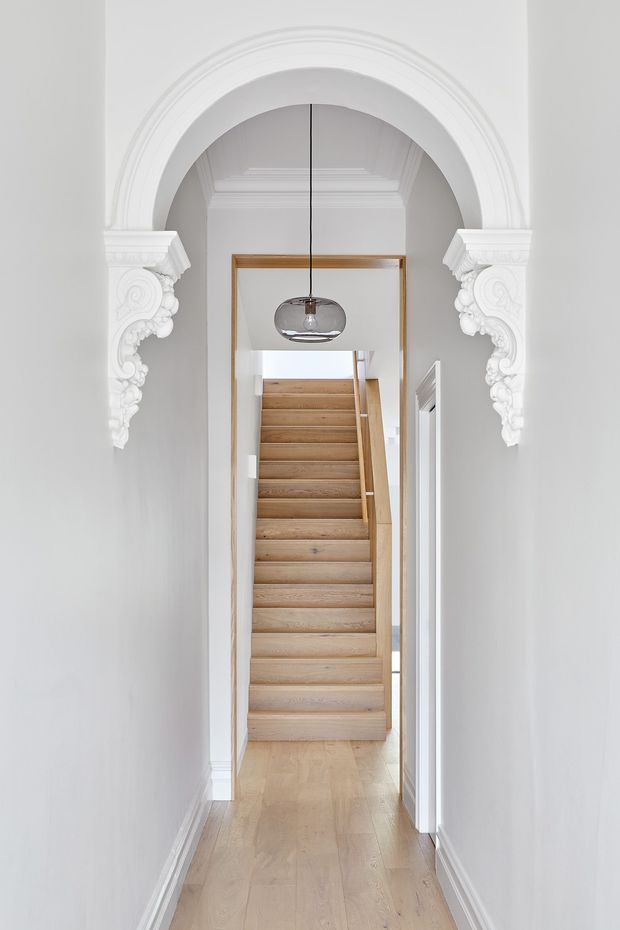 A bright, narrow residential hallway framed by an ornate white arched opening with elaborate plaster corbels and moulded detailing. The corridor has pale timber flooring, white walls and trim, a timber-framed doorway, and a light timber staircase rising at the far end. A suspended glass pendant hangs centrally beneath the arch.