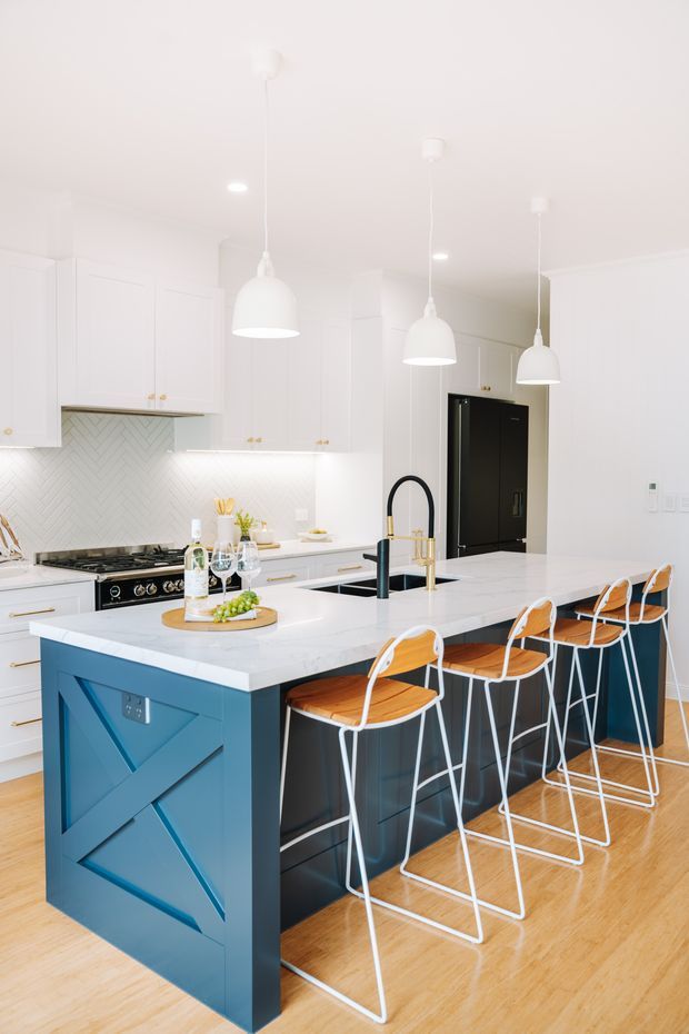 Bright, contemporary white kitchen with a large island featuring deep blue panelled cabinetry and a white benchtop. The island includes a black-and-brass faucet and seating for several people on white wire-frame bar stools with timber seats. White shaker-style cabinetry, a herringbone tiled splashback, integrated black refrigerator, pendant lights, and light timber flooring complete the space.