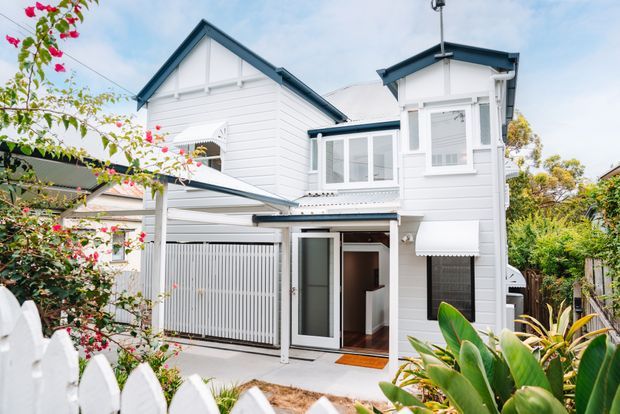 Exterior view of a bright white weatherboard home with a steep gabled roof, projecting upper-level bay, covered entry, and a covered carport or parking structure. The house is framed by a white picket fence and lush garden planting.