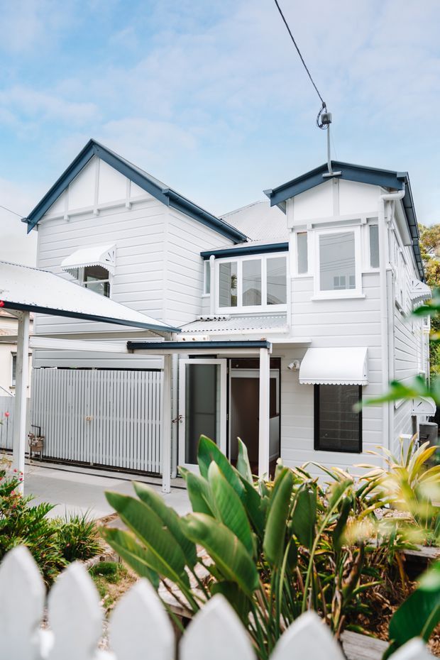 Exterior view of a two-storey white weatherboard house with prominent gabled roof forms, decorative window awnings, a covered carport or entry structure, and a landscaped front garden behind a white picket fence. The building has a bright, traditional residential character with dark blue-grey roof trim and corrugated metal roofing.