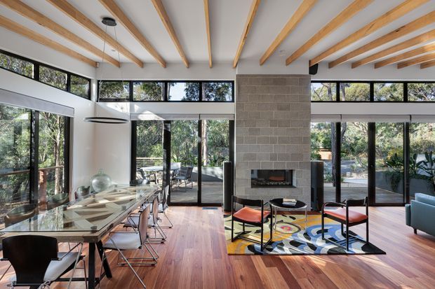Open-plan mid-century-inspired interior combining a dining area and living room, with extensive black-framed glazing, exposed timber ceiling beams, warm timber flooring, and a central grey masonry fireplace. A glass dining table with contemporary chairs occupies the foreground, while colourful lounge chairs and a blue sofa sit around the fireplace.