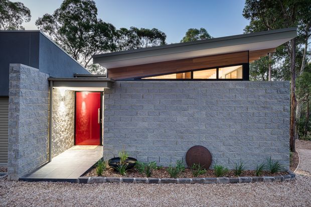 Single-storey contemporary house exterior in a wooded setting, featuring a low-pitched skillion roof, dark cladding, exposed timber soffits, a grey concrete-block privacy wall, and a vivid red entry door. The facade is photographed from the gravel approach with sparse native-style planting and warm interior light visible through high clerestory glazing.