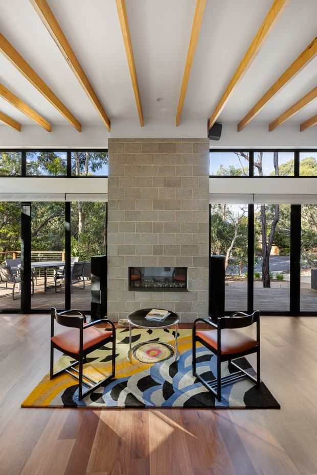 A bright, double-height living room with extensive black-framed glazing opening to a timber deck and wooded landscape. A central exposed concrete-block fireplace wall anchors the room, while exposed timber ceiling beams, warm timber flooring, a colourful geometric rug, and two black-framed leather lounge chairs create a refined mid-century-inspired interior.