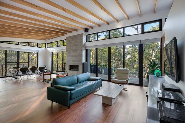 A bright, open-plan living room with a dining area, floor-to-ceiling glazing, and a prominent stone fireplace. Exposed timber ceiling beams span the room, while warm timber flooring contrasts with the dark-framed windows and teal sofa. The space opens visually to a leafy outdoor setting and includes a contemporary media wall and coffee table.