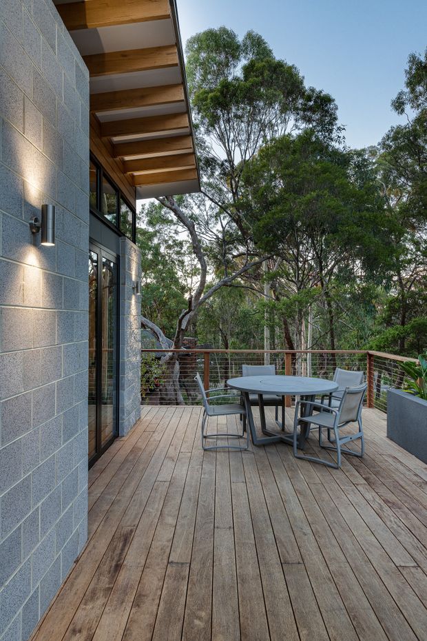 A timber-clad outdoor deck adjoining a contemporary home, with a grey masonry wall, black-framed glazing, exposed timber roof beams and a metal cable balustrade. A round outdoor dining table and chairs sit among mature trees, creating a private bushland setting.