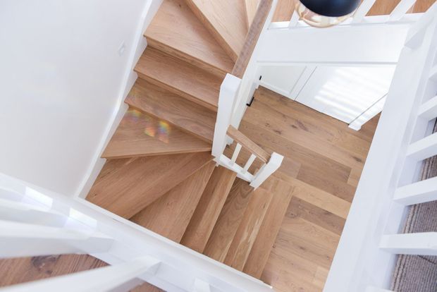 Top-down view of a bright residential stairway with white-painted balustrades and trim, warm natural timber stair treads and matching timber flooring. The stair turns through a compact landing, with strong daylight entering from nearby windows or glazing.