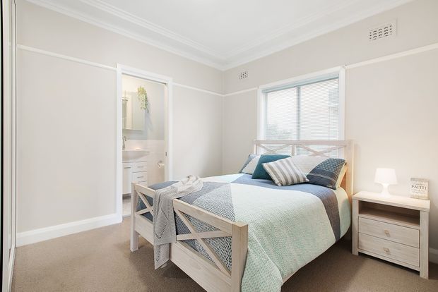 Bright, neatly styled bedroom with a light timber bed, layered blue and white bedding, coordinating cushions, a small bedside table, carpeted floor, and a window with white blinds. A bathroom is visible through the open doorway.
