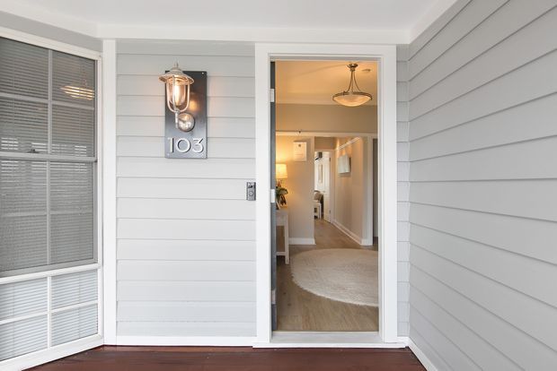 Exterior view of a residential entry porch with grey horizontal weatherboard cladding, white trim, a glazed front door, wall-mounted lantern, house number plaque, and a glimpse of the warm-toned hallway and pendant light inside.