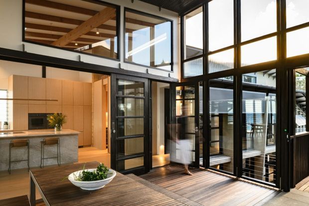 A bright, contemporary double-height interior centered on a kitchen, dining area, and expansive black-framed glazing. Warm timber floors, exposed timber ceiling beams, pale wood cabinetry, and a stone island create a refined modern atmosphere. A glazed upper-level mezzanine and open stair or landing are visible, with a timber outdoor deck beyond the glazing.