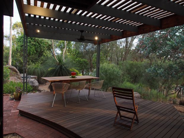A covered outdoor dining deck surrounded by dense native vegetation. The dark timber decking supports a timber dining table with modern chairs and a folding timber lounge chair. Above, a substantial timber pergola with regularly spaced slatted rafters provides filtered light, with integrated warm lighting and a ceiling fan. Brick paving and large natural rocks edge the deck, creating a secluded garden setting.