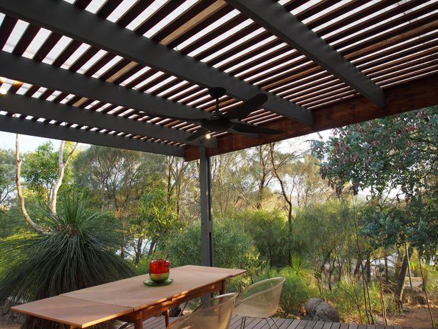 Outdoor dining and entertaining area beneath a broad pergola with dark structural beams, closely spaced timber slats, and an integrated ceiling fan. A timber dining table and transparent chairs sit on a timber deck, overlooking dense native bushland and landscaped vegetation.