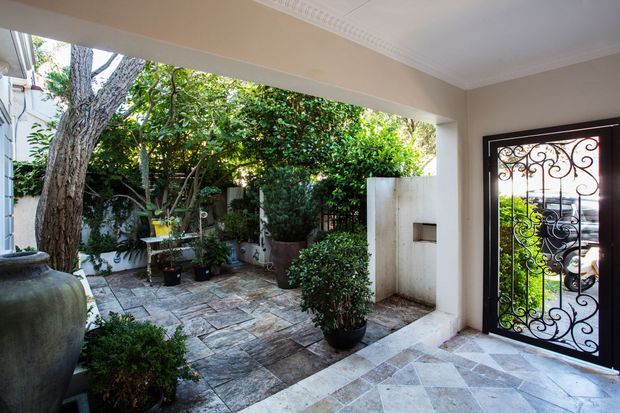 Covered entry foyer opening onto a leafy paved courtyard, with a decorative wrought-iron glazed security door, pale tiled flooring, rendered walls, potted plants, and a large ceramic urn. Traditional detailing includes ornate ceiling cornices and patterned metalwork.