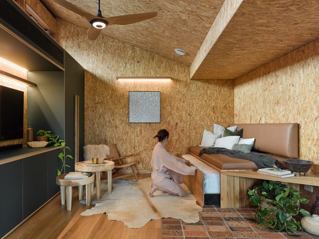 Warm, compact bedroom with OSB wood-panelled walls and ceiling, integrated dark built-in cabinetry and media shelving, a ceiling fan, upholstered bedhead, timber platform bed, and a small seating area. The room combines layered timber textures with soft neutral furnishings, plants, and subdued integrated lighting.