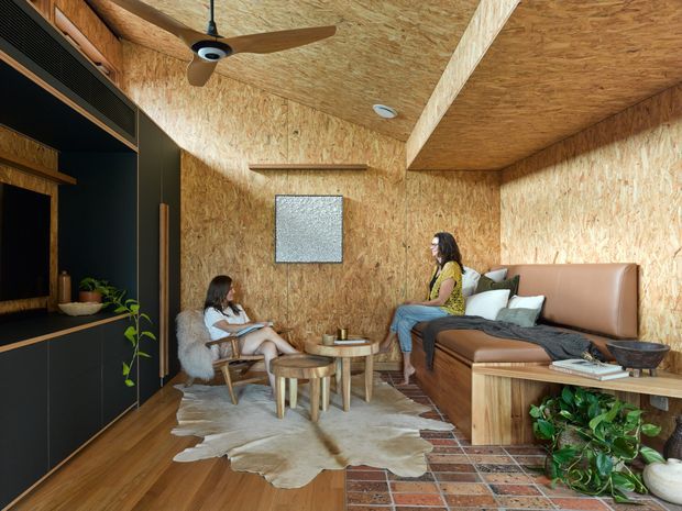 A compact, richly textured living room lined with exposed OSB wood panels across the walls and ceiling. The room includes a built-in dark entertainment unit, a tan leather sectional sofa, timber coffee tables, a cowhide rug, exposed brick flooring accents, indoor plants, and a ceiling fan. Two people are seated in the space, which has a warm, informal, rustic-industrial atmosphere.