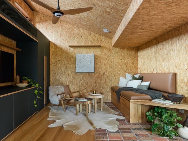 A compact, warmly finished living room with OSB wood-panelled walls and ceiling, integrated dark built-in joinery on the left, a brown leather sofa, lounge chair, nesting coffee tables, and layered rugs. A ceiling fan and recessed lighting provide functional detailing, while plants and soft furnishings add a relaxed atmosphere. The room has a strong rustic-industrial character with exposed wood-chip board surfaces and timber flooring.
