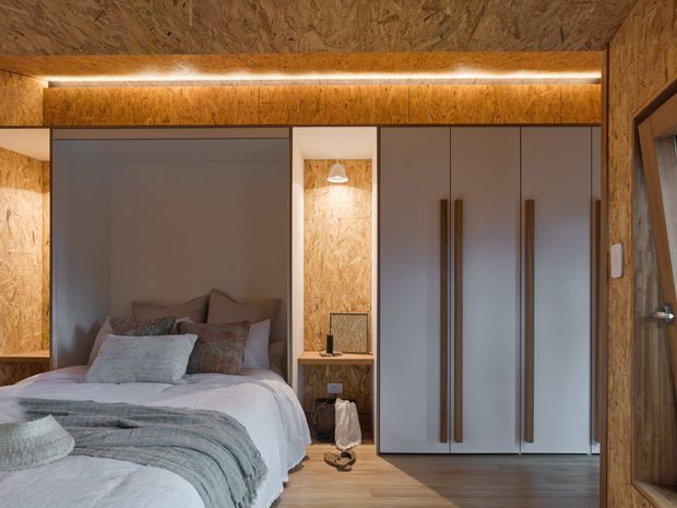 Compact bedroom interior with a bed, built-in wardrobes, bedside niche, and timber OSB wall and ceiling lining. Warm integrated strip lighting contrasts with muted grey cabinetry and soft blue-grey bedding, creating a cozy contemporary cabin-like atmosphere.