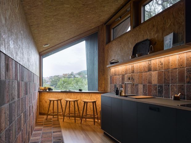 Compact, narrow kitchen with a dark built-in cabinetry run, integrated benchtop and sink, open shelving with under-shelf lighting, and a timber-framed picture window overlooking greenery. The room has a highly tactile, rustic-industrial character, combining exposed oriented strand board on the walls and ceiling with dark, varied-format tiled wall and floor surfaces. A timber breakfast ledge with three stools sits beneath the window.