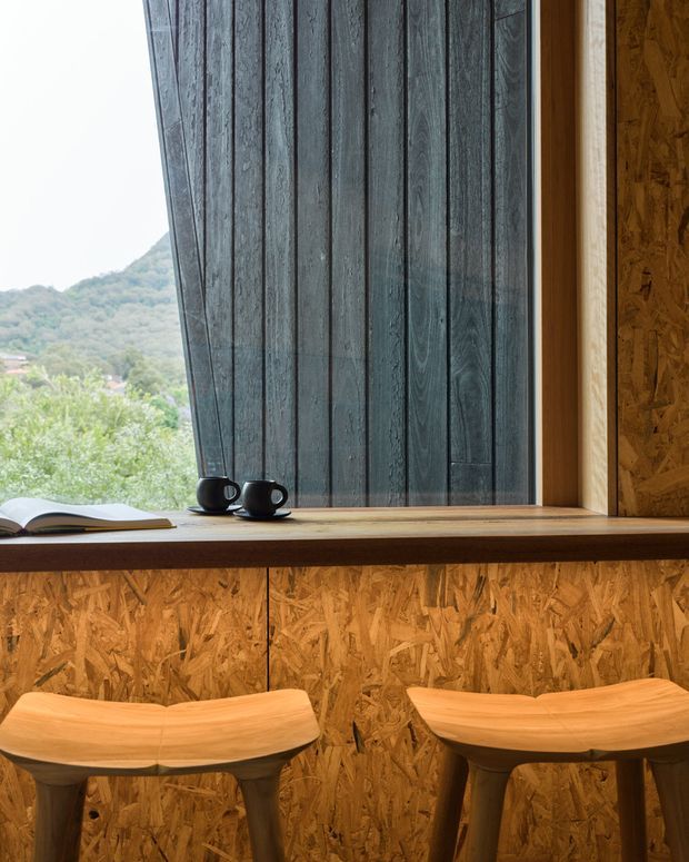 A warm, compact timber-lined interior nook with a broad wooden window ledge functioning as a desk or counter. Two sculptural wooden stools sit below it, while dark vertical timber screening covers the window opening. A book and two black cups rest on the ledge, and a wooded hillside is visible outside.