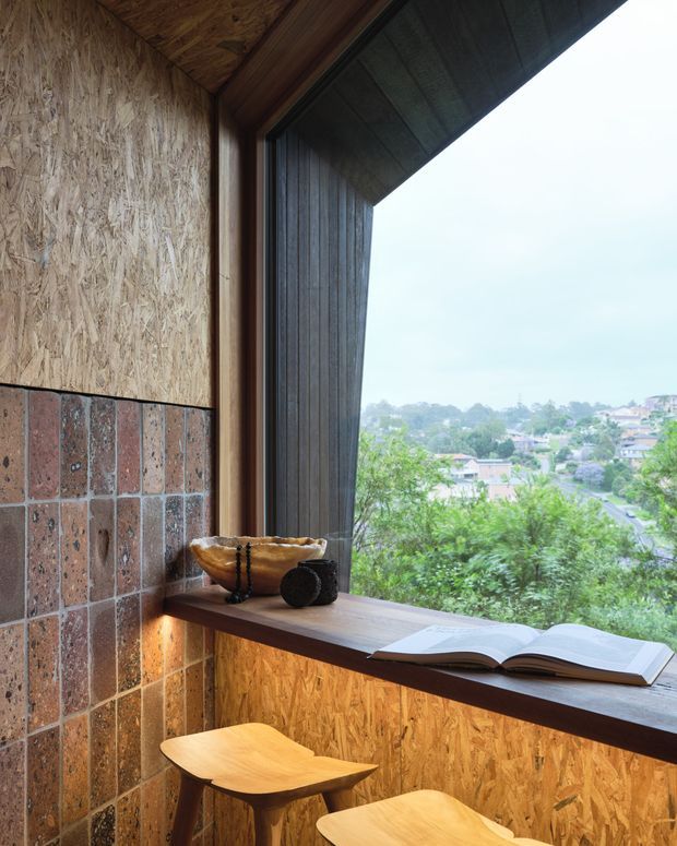 A compact timber-lined reading nook beside a large angled window, with exposed oriented strand board walls and ceiling, a rust-toned brick tile lower wall, dark timber window framing and sill, and two small wooden stools. An open book rests on the deep window ledge, overlooking a leafy suburban landscape.