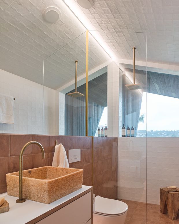 A contemporary bathroom with a walk-in glass shower, wall-mounted rainfall showerheads, freestanding vessel basin, toilet, large mirrored wall, and a warm terracotta-toned tiled dado. The space has pale textured ceiling and wall tiles, brass-toned fixtures, white vanity joinery, and natural light through a large window.