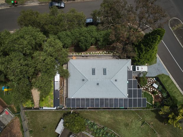 Aerial, top-down view of a suburban residential property. The main house has a large light-grey hipped roof with several skylights and extensive dark solar-panel arrays along the lower roof edges. Mature trees, landscaped garden beds, lawn, paved paths, neighboring streets, and parked vehicles surround the property.