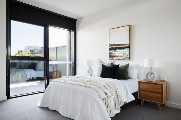 Bright contemporary bedroom with a neatly made bed, white textured bedding, dark accent cushions, timber bedside tables, sculptural lamps, and a framed abstract artwork. Full-height black-framed glazing and a sliding glass door open toward a balcony or exterior view, while grey carpet and white walls create a calm neutral setting.