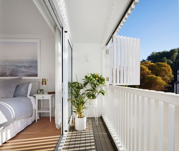 Bright covered balcony or terrace adjoining a bedroom through fully glazed sliding doors. The space has white-painted vertical balustrades and privacy screening, dark timber-look decking, potted greenery, and strong linear sunlight and shadow. A simply styled bedroom with soft grey bedding is visible inside.