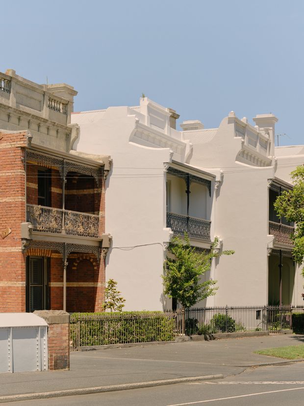 Street-level view of attached heritage terrace houses in bright daylight. The row features ornate wrought-iron balconies and verandah detailing, rendered white facades, decorative parapets and cornices, and an adjacent exposed red-brick terrace. A low iron fence, paved footpath, and residential street occupy the foreground.