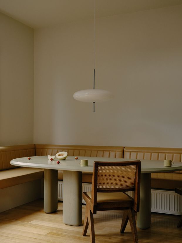 A warm, softly lit dining nook with a built-in caramel leather banquette wrapping around the corner, a pale oval dining table on cylindrical pedestal legs, and a single dark wood chair with a woven cane back. A minimalist white pendant hangs above the table. Neutral plaster walls, timber flooring, and visible radiators complete the understated interior.