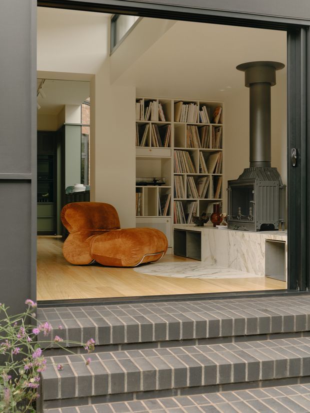 View through a large glazed opening into a refined living room. The room features a sculptural burnt-orange lounge chair, pale timber flooring, extensive built-in shelving filled with books and records, and a dark freestanding wood-burning stove set on a veined marble-clad hearth. Dark grey tiled exterior steps and a small flowering plant frame the foreground.