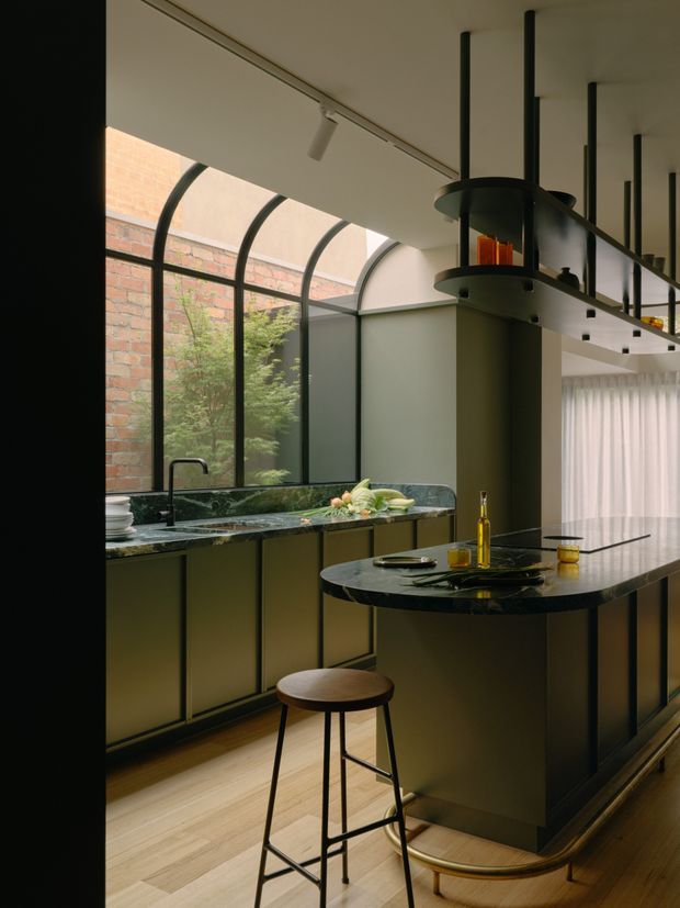 A sophisticated contemporary kitchen with dark green-black fixed cabinetry, polished dark stone benchtops, and a central island with curved edges. The room features a dramatic curved glazed roof or clerestory enclosure with black metal framing, exposed brick visible beyond the glazing, track lighting, and a sculptural suspended black rangehood or fireplace-like feature above the island. Pale timber flooring and sheer curtains soften the predominantly dark, refined palette.