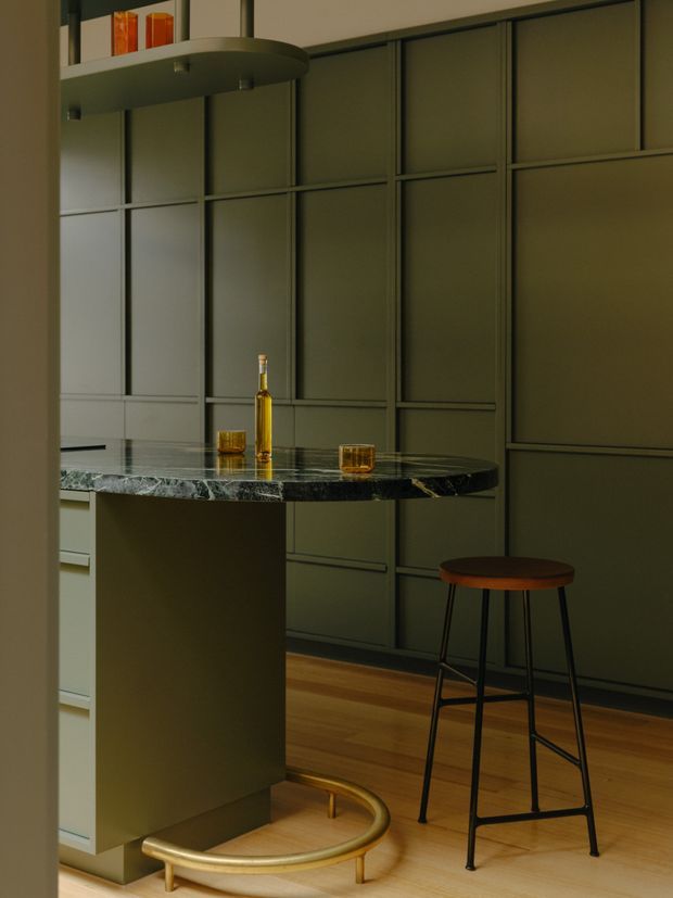A moody, refined kitchen interior with dark olive-green full-height panelled cabinetry, a polished dark veined stone island benchtop, and warm timber flooring. A curved floating shelf with amber glassware hangs above the island, while a tall bar stool and a sculptural brass-and-wood base detail add character. The space has integrated joinery and a restrained, sophisticated palette.