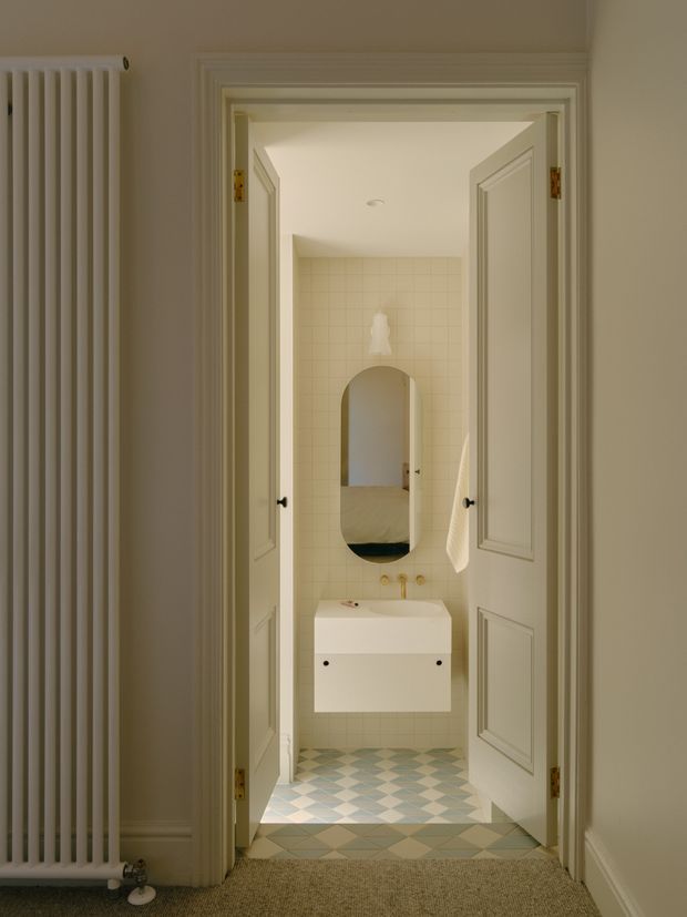 A refined bathroom viewed through open, paneled double doors from a hallway. The compact room features a wall-mounted white basin with brass fittings, an arched mirror, pale square wall tiles, and a patterned blue, grey, and cream tiled floor. A tall white radiator and carpeted hallway are visible in the foreground. The space has a soft, warm, traditional feel with crisp architectural detailing.