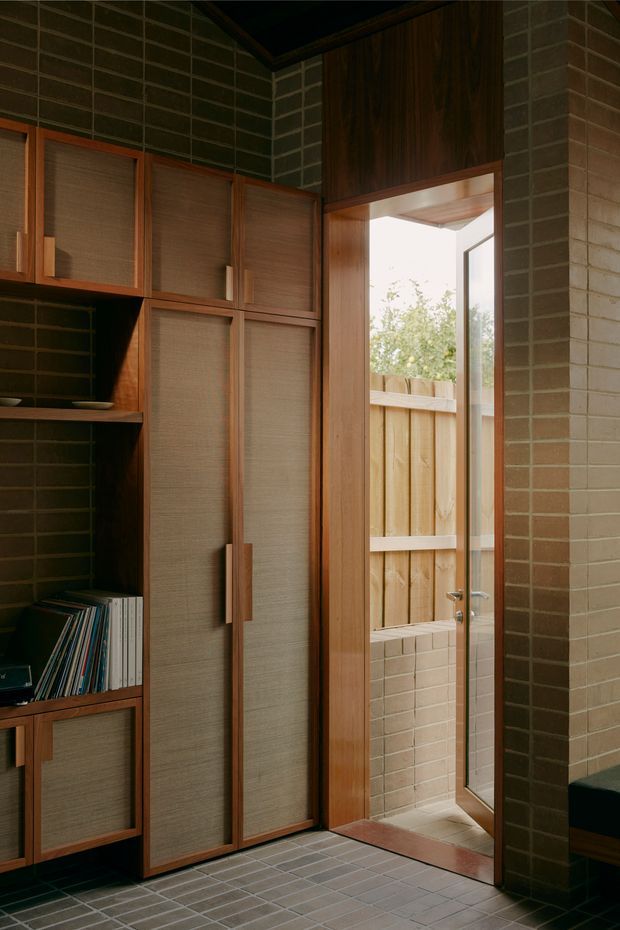 A compact, warmly lit interior passage or entry area with floor-to-ceiling built-in timber cabinetry featuring woven or textured inset panels and integrated handles. Dark green-grey brick walls and matching small-format tiled flooring frame a glazed timber-framed door opening to a small fenced exterior lightwell or courtyard. The space has a rich, tactile mid-century-inspired material palette and strong vertical joinery.