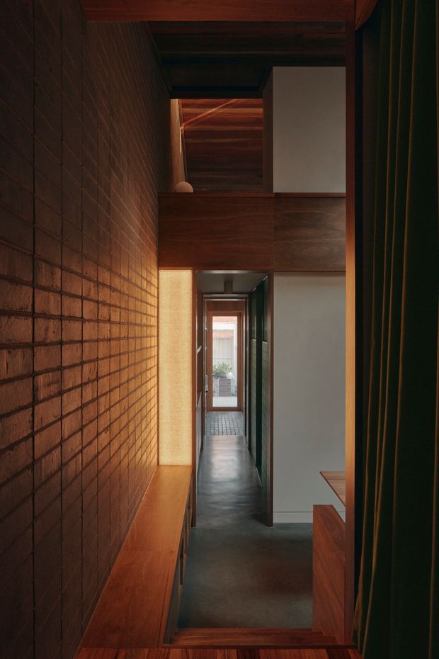 A narrow, moody interior hallway with a richly textured exposed brick wall on the left, dark timber ceiling and joinery, polished dark concrete flooring, and a long timber bench. White plaster walls and a series of aligned doorways lead toward a glazed opening at the far end. Deep green vertical curtains frame the right side, while warm natural and artificial light creates a dramatic, intimate atmosphere.