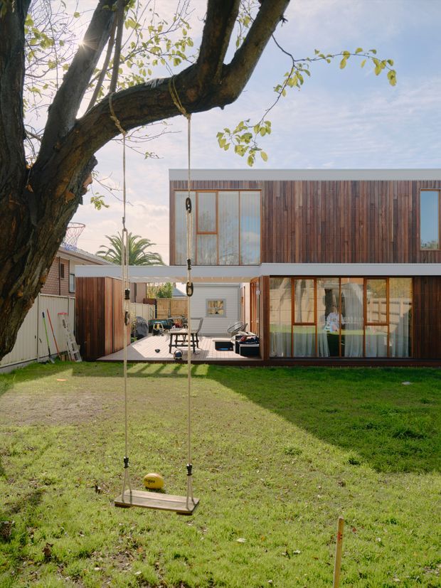 Exterior view of a contemporary two-storey residence with extensive vertical timber cladding, large glazed openings, and a covered outdoor patio. A mature tree with a hanging swing dominates the foreground, with a lawn and small courtyard visible around the house.