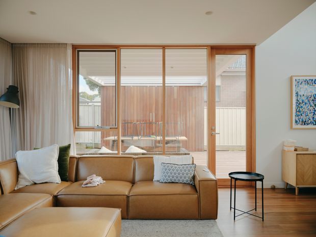 A warm, contemporary living room with a large tan leather sectional sofa and ottoman, floor-to-ceiling timber-framed glazing opening onto a covered timber courtyard or deck. Sheer curtains, a black floor lamp and side table, pale walls, timber flooring, and a small timber sideboard create a refined, inviting interior. A colourful artwork provides a small accent.