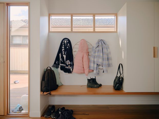 A bright, compact entryway with a built-in timber bench and open shoe storage beneath. Coats, scarves, bags and boots hang or sit on the joinery, while warm timber-framed glazing brings in daylight and overlooks a neighbouring deck. The space has white walls, dark timber flooring and simple contemporary detailing.