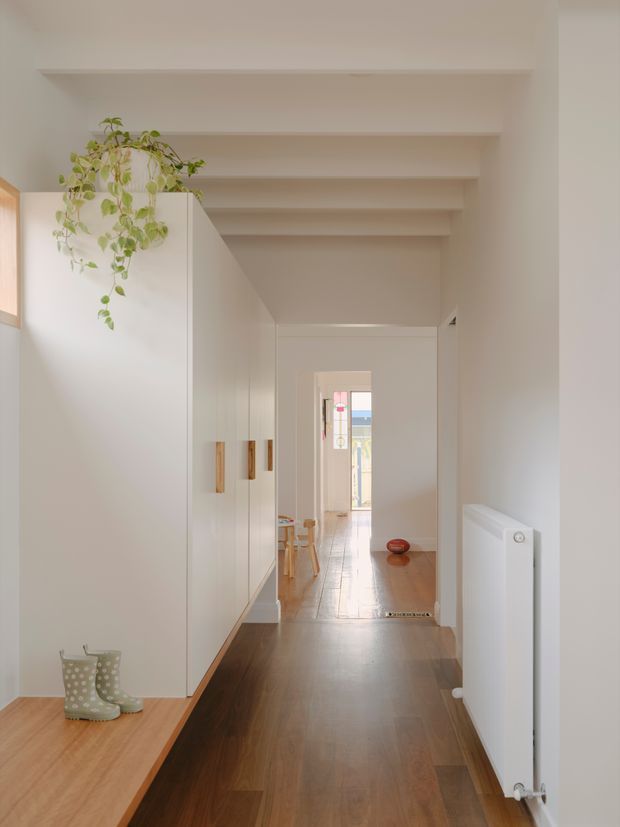 A bright, narrow residential hallway with warm timber flooring and a long run of white built-in cabinetry featuring timber handles. Exposed white ceiling beams create a linear perspective toward a brighter room beyond. A timber bench with small boots sits in the foreground, while a trailing indoor plant adds greenery. The space has a clean, calm, minimalist presentation with warm natural light.