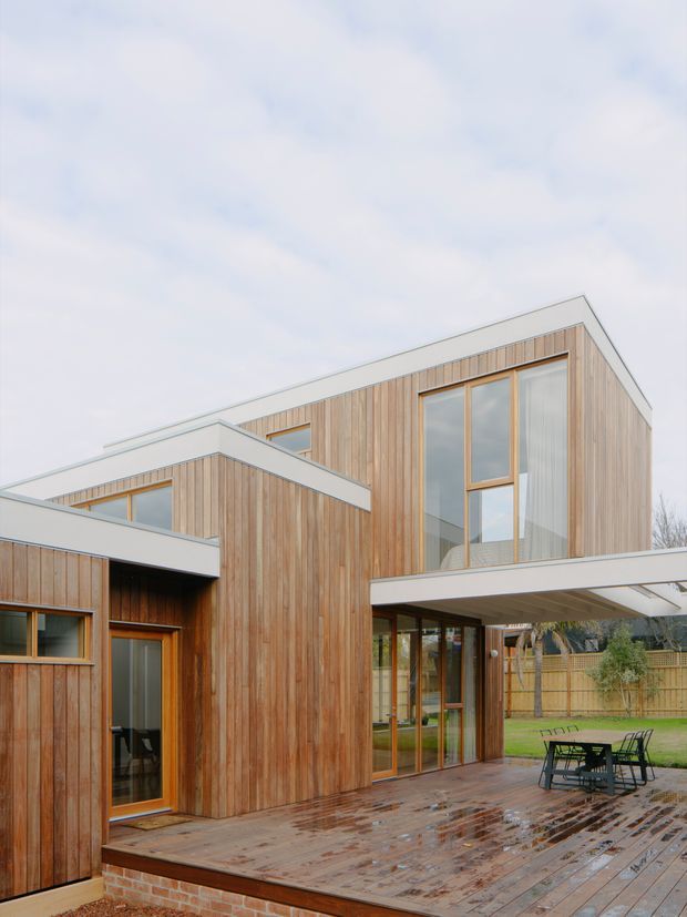 Exterior view of a contemporary two-storey house with warm vertical timber cladding, large timber-framed windows, and intersecting flat-roof volumes. A covered outdoor terrace with timber decking extends from the glazed ground floor, with a fenced garden and outdoor dining furniture visible in the foreground.