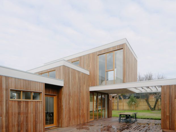 Exterior view of a contemporary multi-volume house arranged around a covered outdoor courtyard. The building features extensive vertical timber cladding, large timber-framed windows, flat roof forms, and a wet brick-paved terrace with outdoor dining furniture. A fenced garden is visible beyond the covered opening.